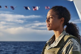 Woman in military uniform looking at the sea, with waving flags of the ship in the background, AI