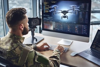 A soldier works in front of a large screen showing technical details and programming of a drone, AI