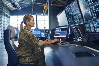 A woman in military uniform works at several computer monitors in a modern control centre, AI