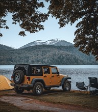 Off-road vehicle parked by a lake with tents and chairs, surrounded by mountain scenery and an