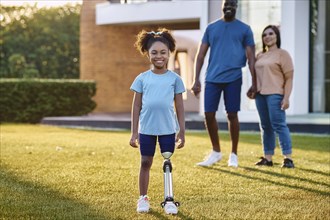 Smiling girl with prosthetic leg in the garden, parents standing smiling in the background next to