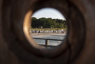 View through a round wooden hole onto a beach with beach chairs and green trees in the background,