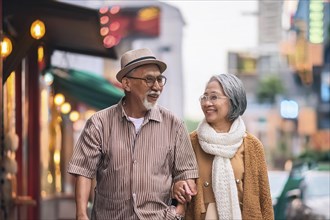 An elderly couple walk hand in hand through an illuminated city street and smile, ai generated, KI