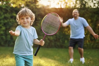 Smiling child in the garden playing with a badminton racket, in the background an adult person, ai