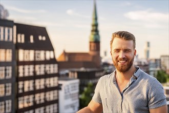 Man with beard enjoying the evening sun on a roof terrace with a view of the city, ai generated, KI