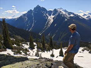 Man standing on a snowy mountain peak surrounded by tall mountains under a clear blue sky. British