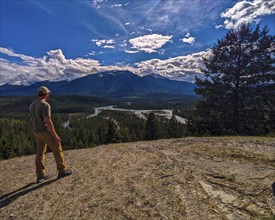 Man standing on a hill looking at a river winding through a forest with mountains in the background