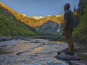 Man standing by a river, watching the mountains illuminated by the setting sun. Montana, United