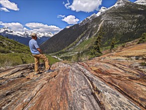 Man standing on rocky terrain with mountains and greenery under a blue sky with clouds. British