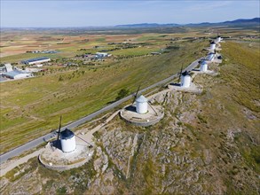 Row of windmills on a grassy hill along a road overlooking wide fields and a village in the