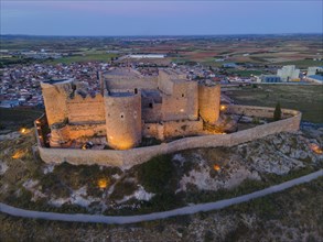 An old castle illuminated at night with a town and wide fields in the background at sunset, aerial