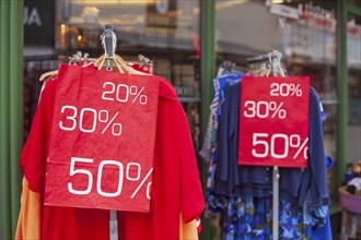 Red sign discount on a shopping bag in front of a clothing rack with t-shirts and shop window,