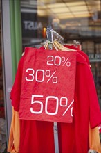 Red sign discount on a shopping bag in front of a clothing rack with t-shirts and shop window,