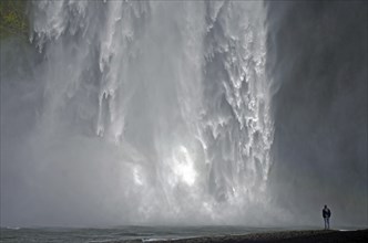 A man stands in front of an impressive waterfall, surrounded by splashing spray, Skogafoss,