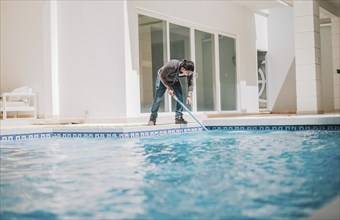 Person cleaning crystal clear pool with special brush. Home pool cleaning with wall brush