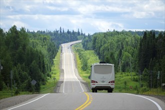 Long road through dense forest, a motorhome driving on it, bright clouds in the sky and a summer