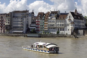 A boat sails along townhouses on the banks of a river under a cloudy sky, Rhine, Basel,