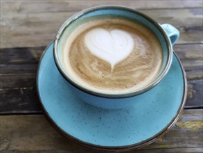 A blue ceramic cup with a heart made of milk foam on a wooden table embodies cosiness