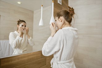 A woman in a white robe is applying skincare mask to her face, standing before a large mirror in a