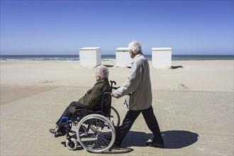 Retired husband taking disabled elderly wife in wheelchair for a walk on promenade along the coast