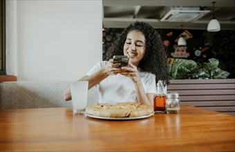 Afro girl taking a picture of a pizza with the phone in a pizzeria. Smiling young woman using phone