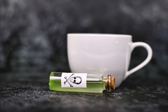 Small vial with cork and green liquid and poison skull label next to tea cup on dark background