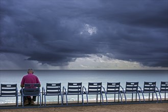 Elderly man sitting on bench at dyke and watching dark, menacing storm clouds over the sea