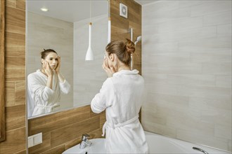 A woman in a white robe is washing her face, standing before a large mirror in a well-lit,