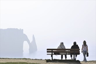 Elderly tourists sitting on bench looking at the Porte D'Aval, a natural arch in the chalk cliffs