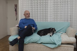 A mature man sits comfortably on the sofa in his home, accompanied by his greyhound. The sofa