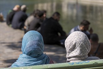 Two woman with headscarves sit on a bench, the men at the back by the old canal, Paris, France,