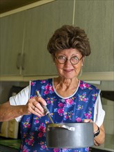 Granny with old smock apron, glasses and wig in the kitchen, holding cooking pot and ladle,