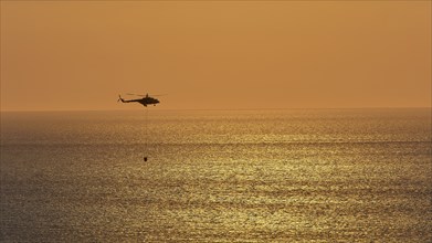 Helicopter, helicopter, fire-fighting helicopter, close over the sea, sunset, sun reflections on