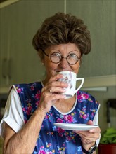 Granny with old smock apron, glasses and wig in the kitchen, holding coffee cup, Germany, Europe