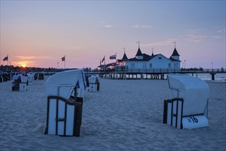 Beach chairs on the sandy beach, people strolling on a pier with buildings in the sunset, clear,