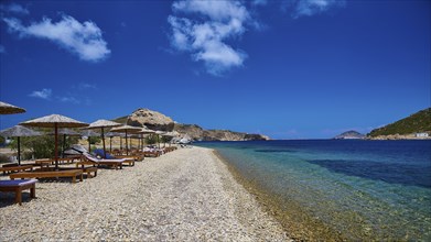 An empty beach with deckchairs and straw umbrellas on the clear blue waters of a sea under a bright