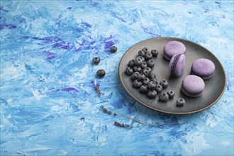 Purple macarons or macaroons cakes with blueberries on ceramic plate on a blue concrete background.