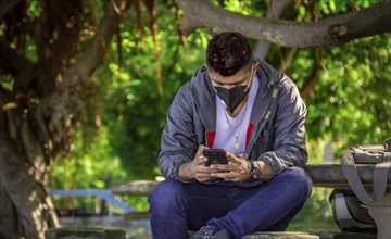 Man with a mask sitting on a bench with his phone, young man with a mask sitting with a cell phone,