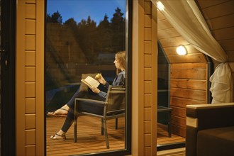 View through the window of a young woman sitting on the terrace in the evening with a book