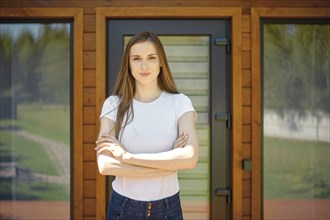 A young woman with long hair stands confidently in front of modern wooden house doors, conveying