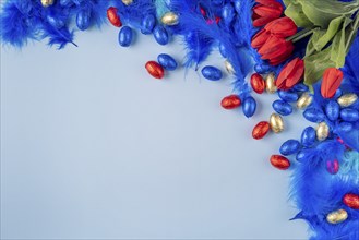 Colourful composition with blue feathers, red tulips and Easter eggs on a blue background