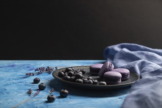 Purple macarons or macaroons cakes with blueberries on ceramic plate on a blue and black background