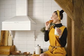 Young woman holds a glass of milk and bites bun. She is sitting on kitchen tabletop in profile