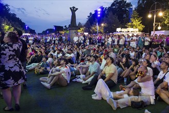 Fans watch the match on one of the rear monitors in front of the statue The Caller in the fan zone