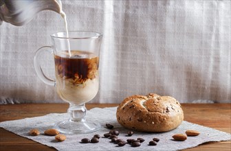 Glass cup of coffee with cream poured over and bun on a wooden background and linen textile. close