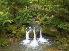 An impressive waterfall in the forest under a bridge with dense green plants and rocks, aerial