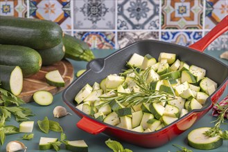 Diced courgettes in a red pan, decorated with fresh herbs in front of colourful tiles and stacked