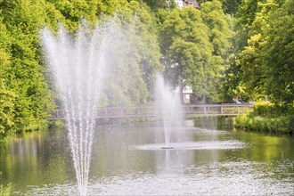 Water fountains bubble in a tranquil lake, surrounded by lush greenery and a bridge path in the