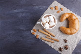 A cup of hot chocolate with marshmallow, croissant and spices on linen napkin and black wooden