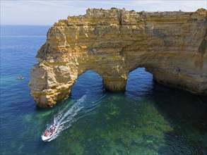 A large boat passes under natural arches made of rocks in crystal clear water on the coast, aerial